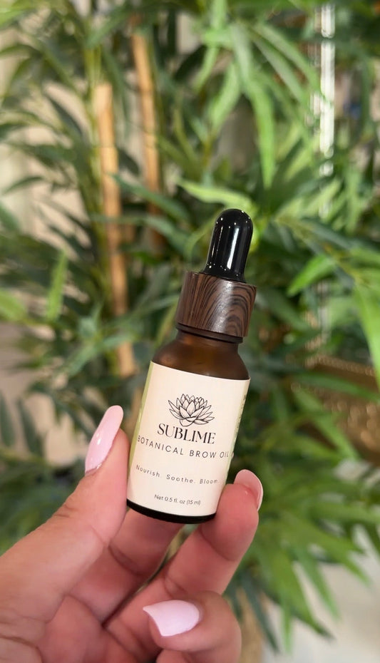 Sublime Botanical Brow Oil in an amber glass dropper bottle with a wood-grain cap, held in a hand in front of green plants